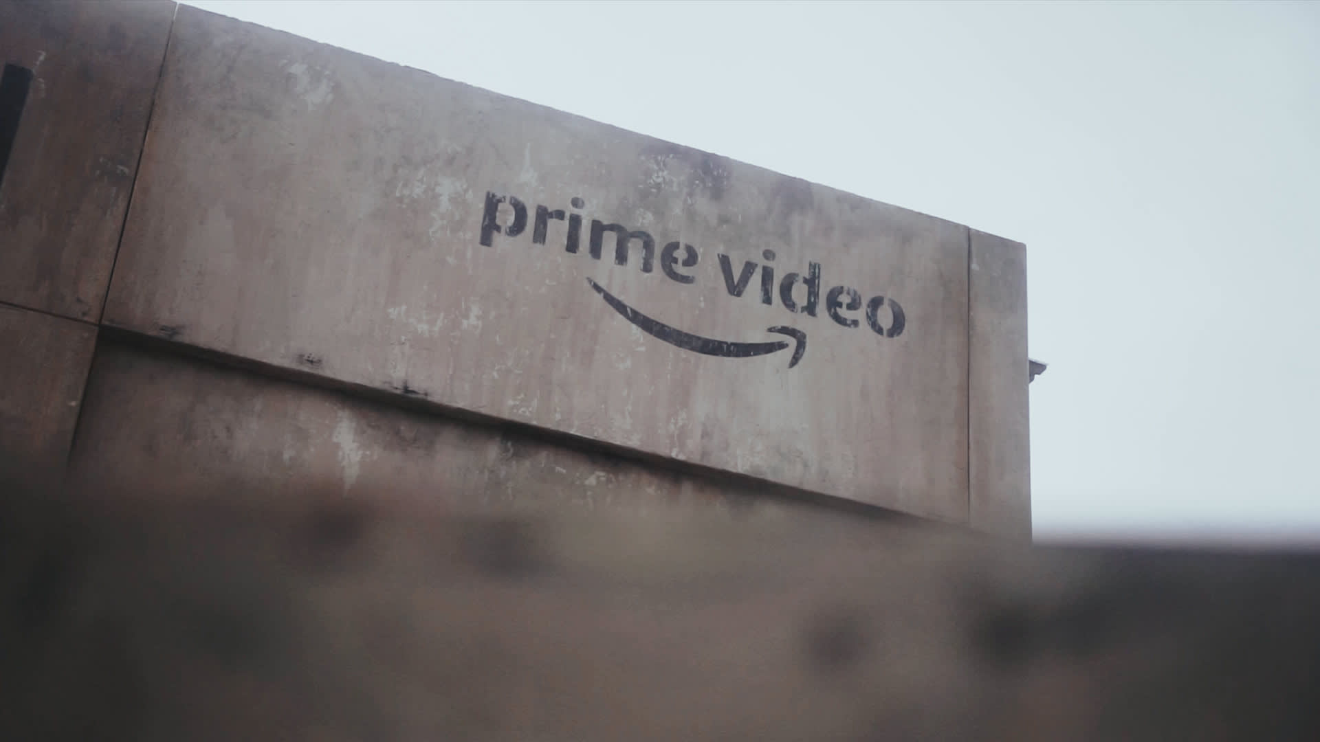 A weathered concrete wall displays the prime video logo with the signature arrow, partially obscured at the bottom, against an overcast sky.