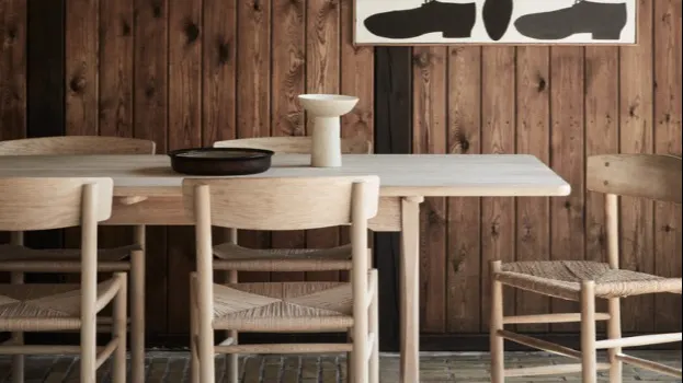 A minimalist dining area with a light wood table, four matching woven-seat chairs, a ceramic vase, a bowl, and a rustic wood-paneled wall featuring abstract black and white artwork.