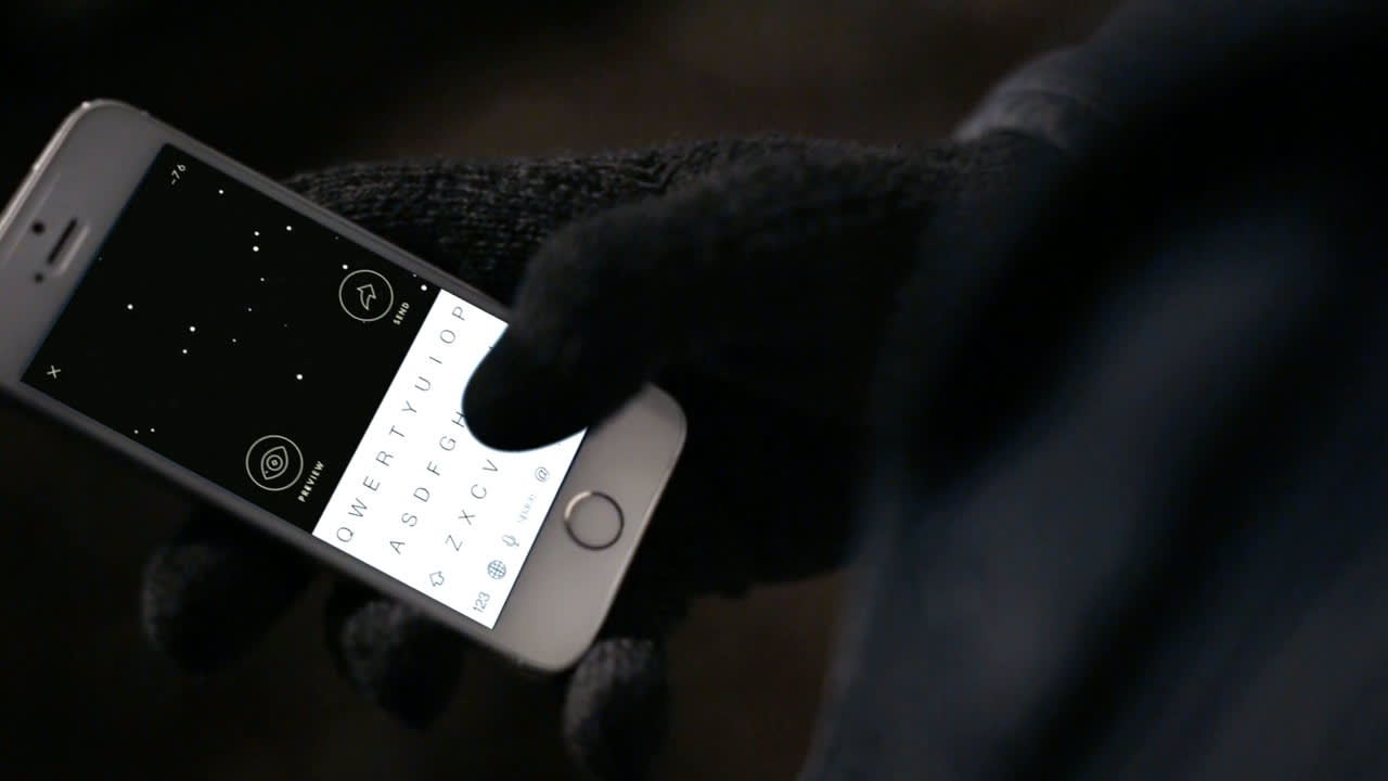 A person wearing black gloves holds a white smartphone, showing a star map app and an on-screen keyboard. The phone is being used in a dark environment.