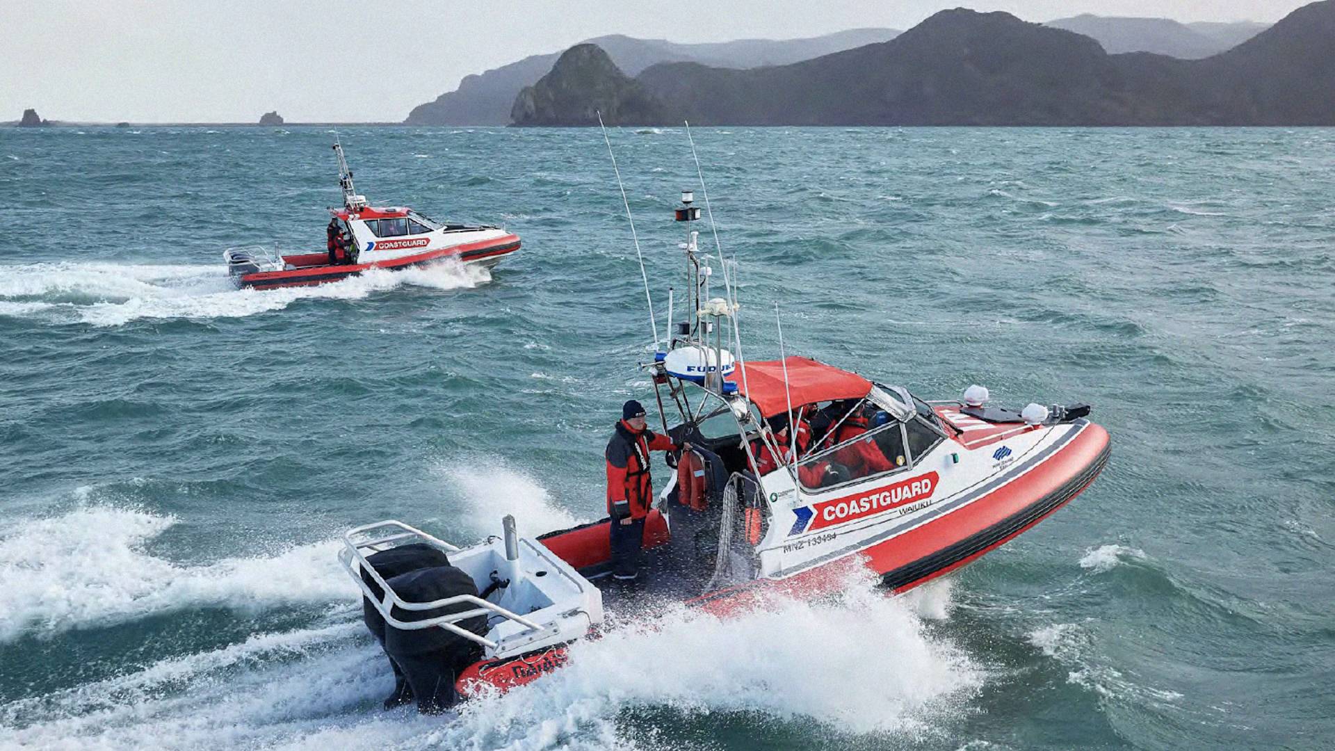 Two red and white Coastguard boats with crew in life jackets speed across choppy water, creating waves, with green hills and a cloudy sky in the background.
