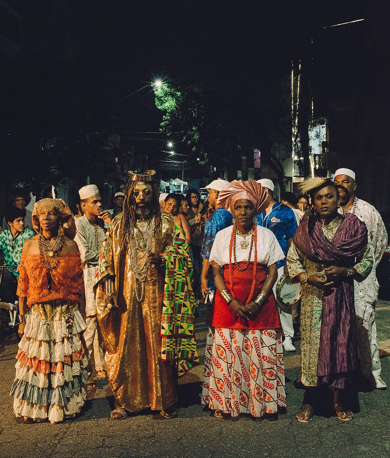 A group of people in vibrant traditional clothing stand in a line on a street at night, participating in a cultural or religious event. The background is dark, with more people gathered behind them.