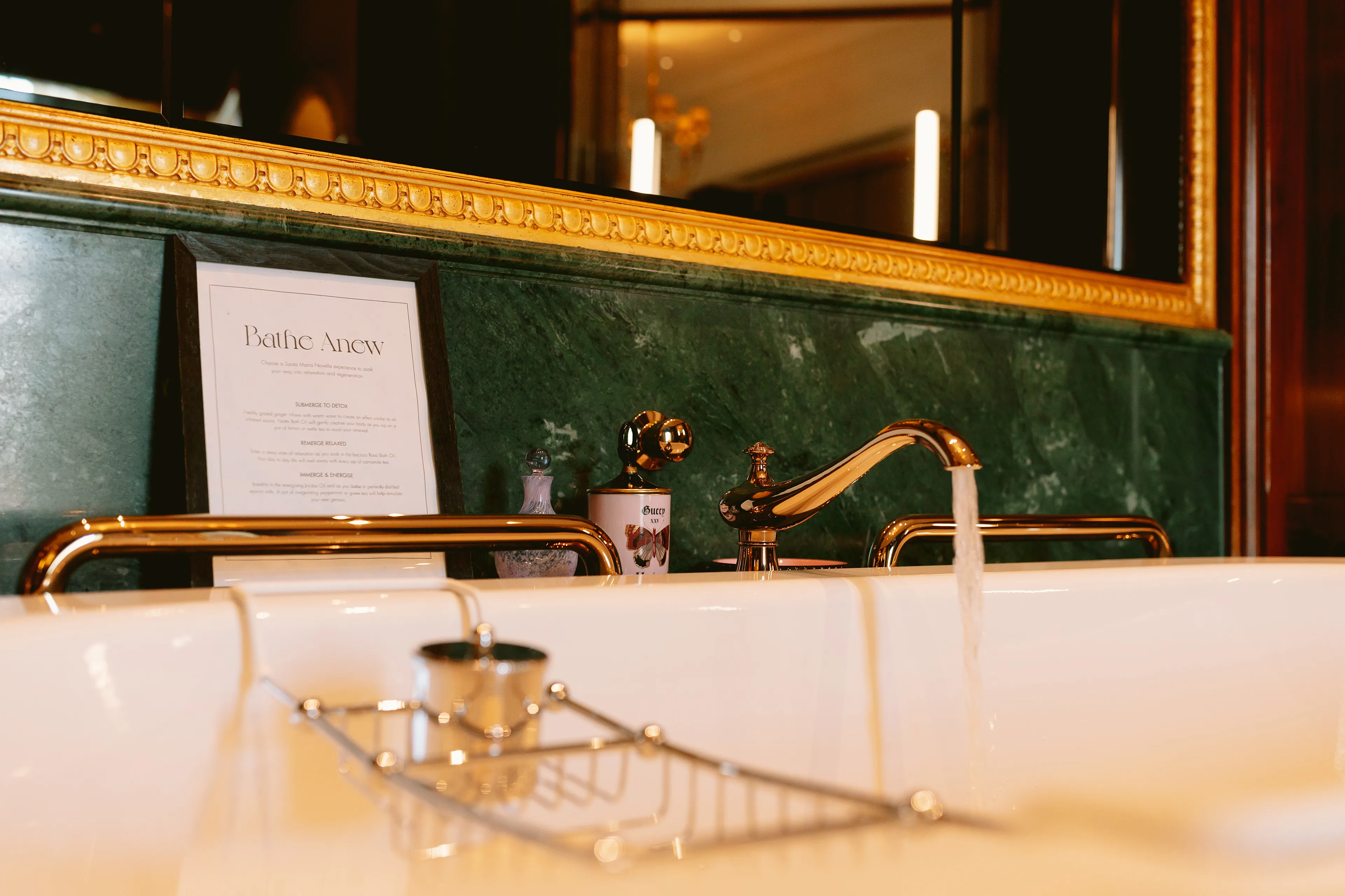 A luxurious bathroom sink with gold fixtures, water running from the tap, and a green marble backsplash. A framed sign and toiletries are on the counter; a metal tray holds items over the white basin. A large mirror is in the background.