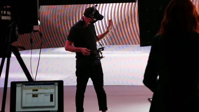 A person wearing a VR headset and holding a controller stands on a stage with colorful, abstract lighting. A computer screen with software is visible in the foreground, and another person stands off to the right.