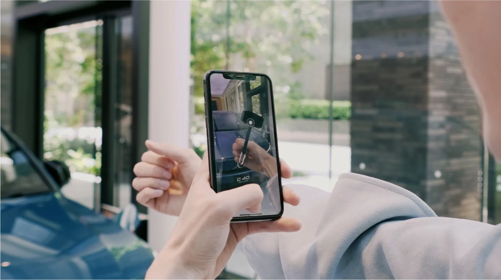 A person holding a smartphone takes a photo of a car key, with a blue car and large windows visible in the background.