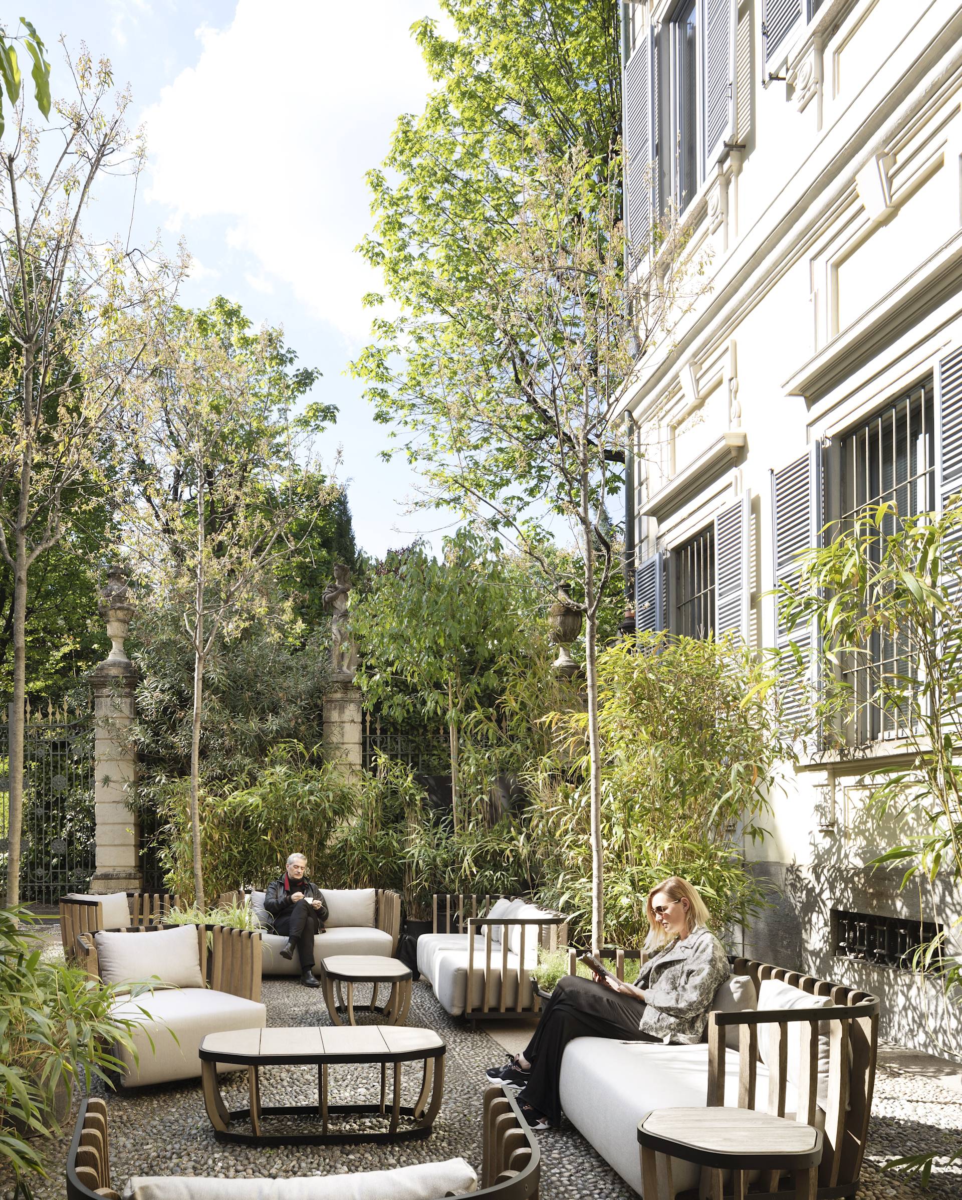 Two people relax in a sunny outdoor patio with modern lounge chairs, surrounded by lush greenery and trees next to a light-colored building with tall windows and shutters.