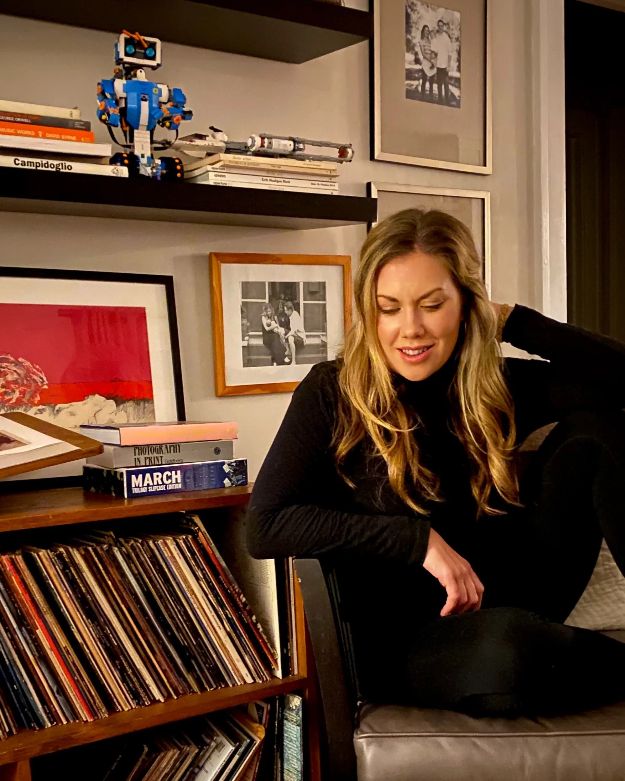 A woman with long blonde hair in a black outfit sits on a chair, smiling with her eyes closed, in a cozy room filled with books, vinyl records, framed photos, and a blue LEGO robot on a shelf.