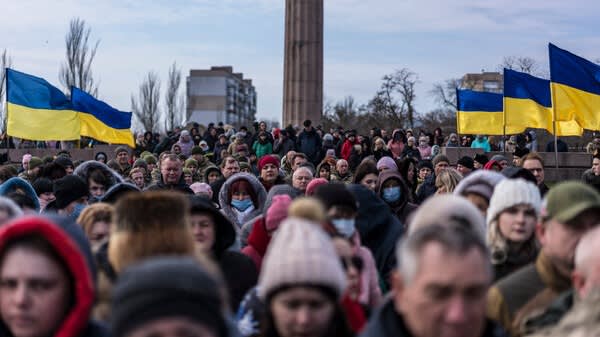 A large crowd of people gathers outdoors, many wearing winter clothing. Several Ukrainian flags are visible in the background, with some buildings and leafless trees behind them. The sky is cloudy.