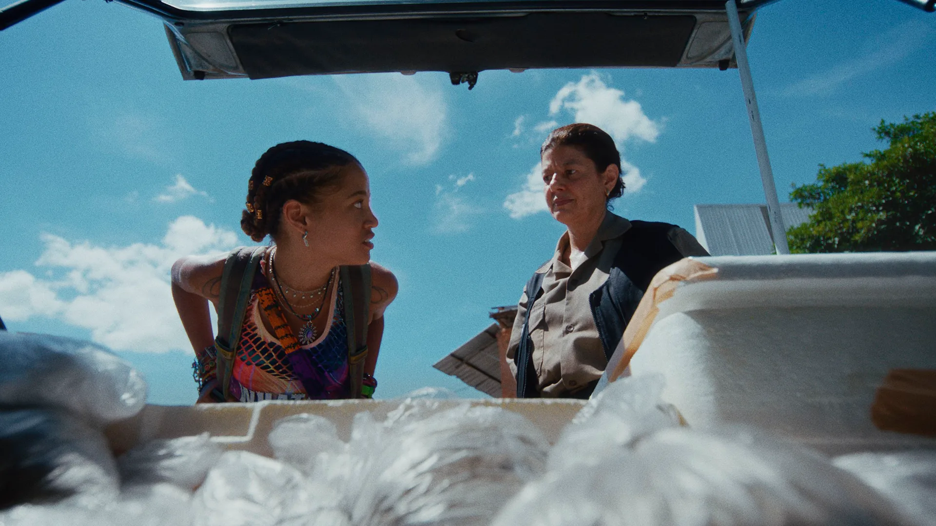 Two women stand outside looking into the open trunk of a car filled with large white bags, under a bright blue sky with scattered clouds. One woman wears colorful clothing and the other wears a uniform.