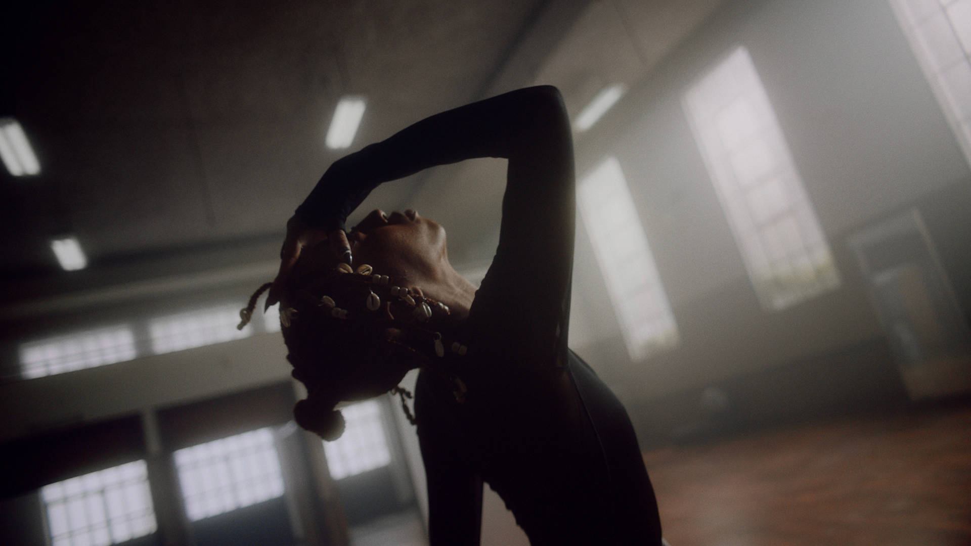 A person in a fitted black outfit leans backward with their hands on their head, dancing in a spacious, sunlit room with large windows and a high ceiling.