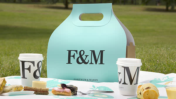 A light blue Fortnum & Mason picnic box sits on grass with two branded coffee cups, pastries, and sandwiches arranged on a white picnic mat in a park setting.
