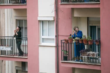 Two women stand on separate apartment balconies, clapping. One is dressed in dark clothing, the other in a blue coat surrounded by potted plants. The buildings have pink and beige walls and white-framed windows.