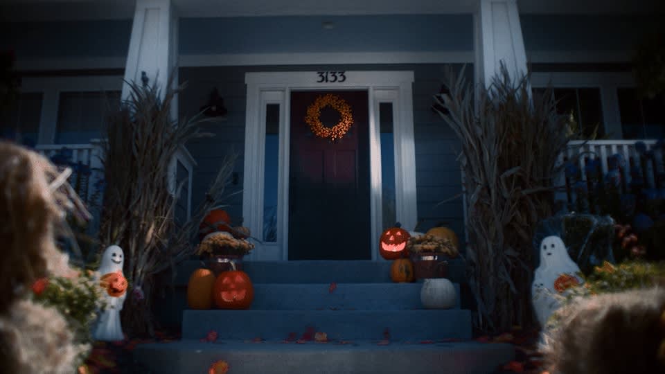 A front porch decorated for Halloween with jack-o’-lanterns, pumpkins, cornstalks, and ghost figures. Orange leaves are scattered on the blue steps, and a wreath hangs on the door with the number 3133 above it.