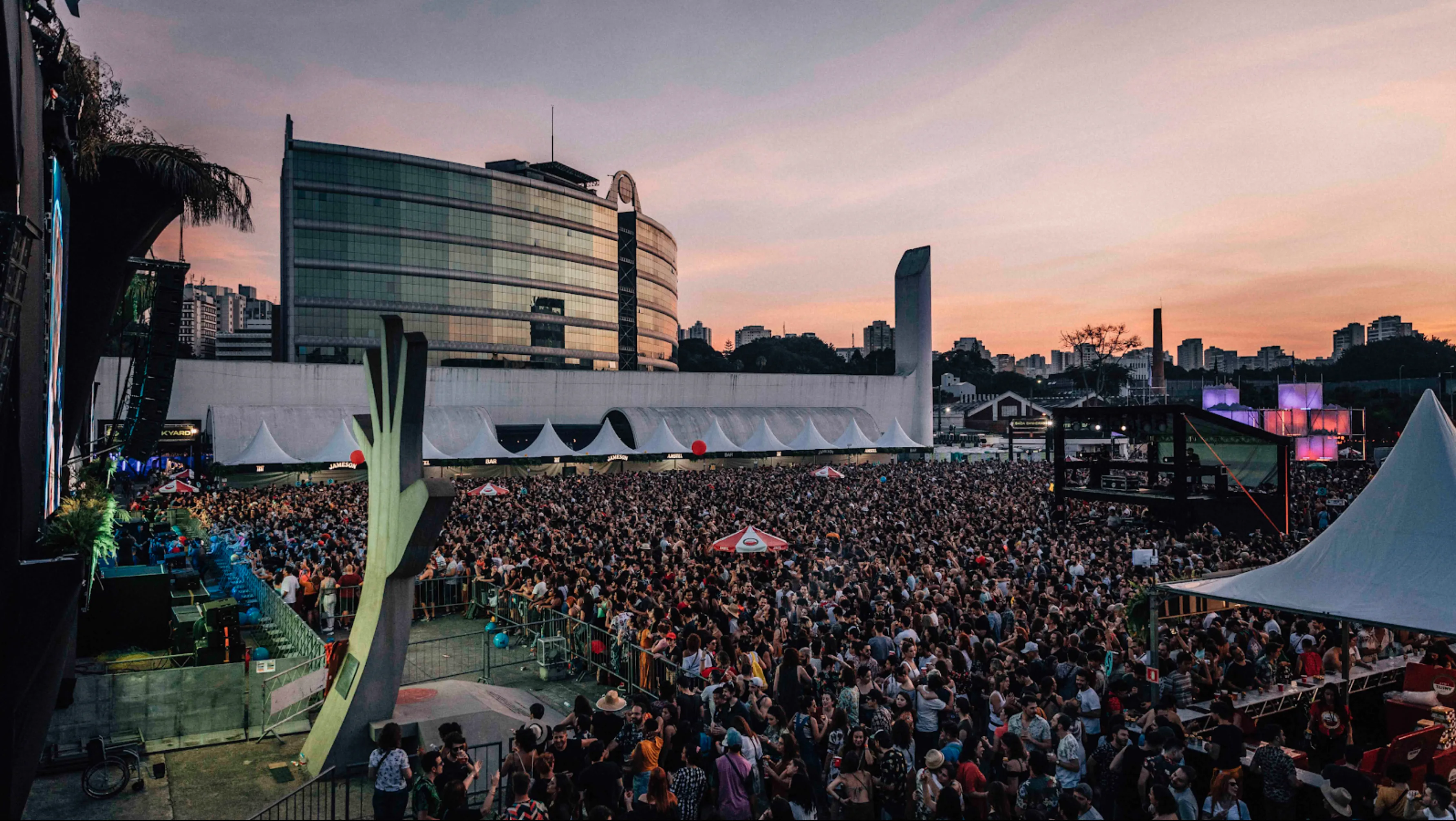 A large crowd gathers at an outdoor music festival at sunset, with a modern, glass-fronted building and city skyline in the background. Tents, lights, and festival booths are visible among the attendees.