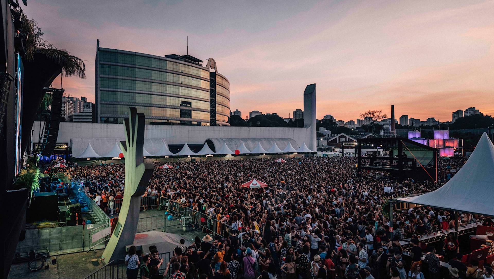 A large crowd gathers at an outdoor music festival at sunset, with a modern, glass-fronted building and city skyline in the background. Tents, lights, and festival booths are visible among the attendees.