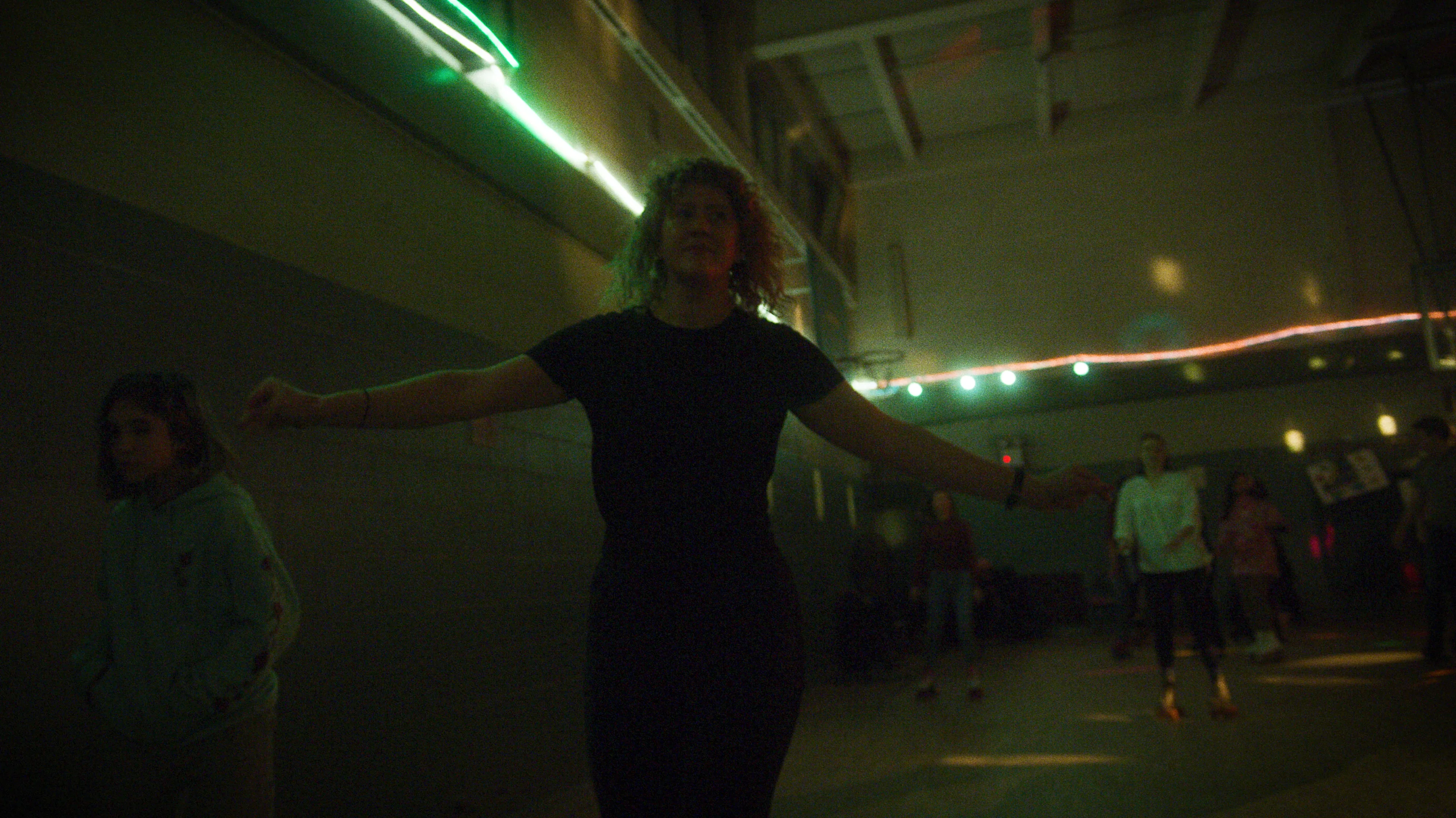 A woman in dark clothing roller skates in a dimly lit gym with neon lights. Other people skate in the background, and a young person in a hoodie stands near the wall. The scene has a moody, retro atmosphere.