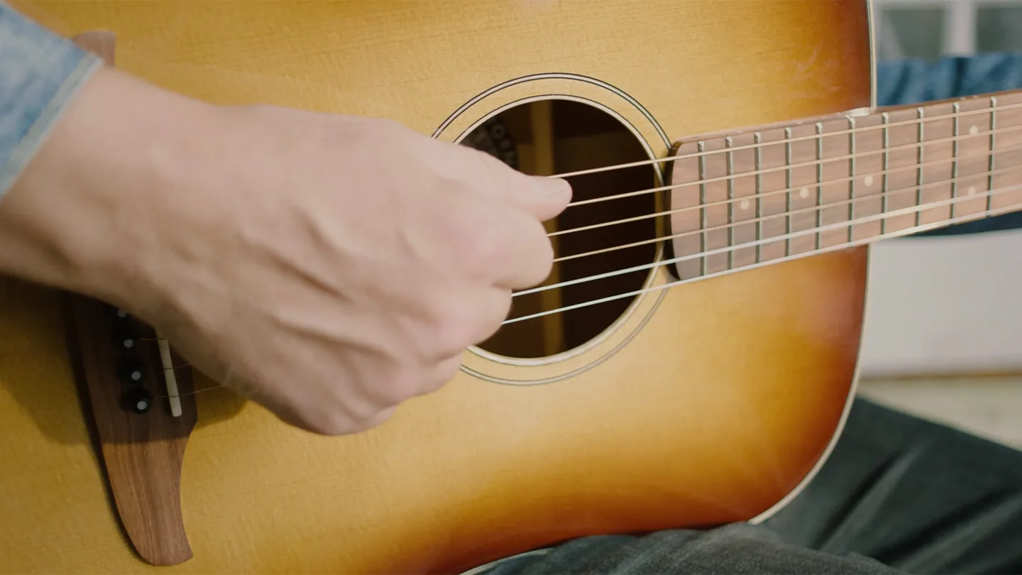 Close-up of a person’s hand strumming the strings of an acoustic guitar, showing the sound hole, bridge, and part of the fretboard. The person is wearing a blue shirt and sitting down.