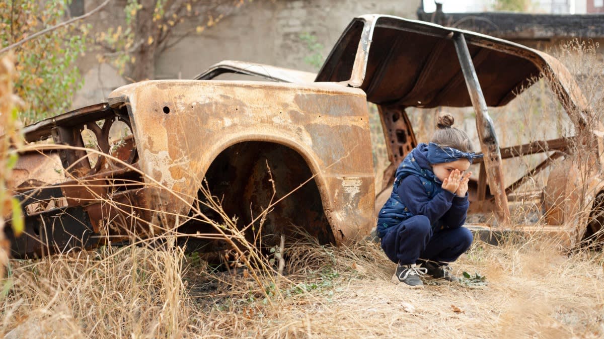 A young girl in a blue outfit crouches and covers her face with her hands next to the rusty remains of an abandoned car in a dry, overgrown field.
