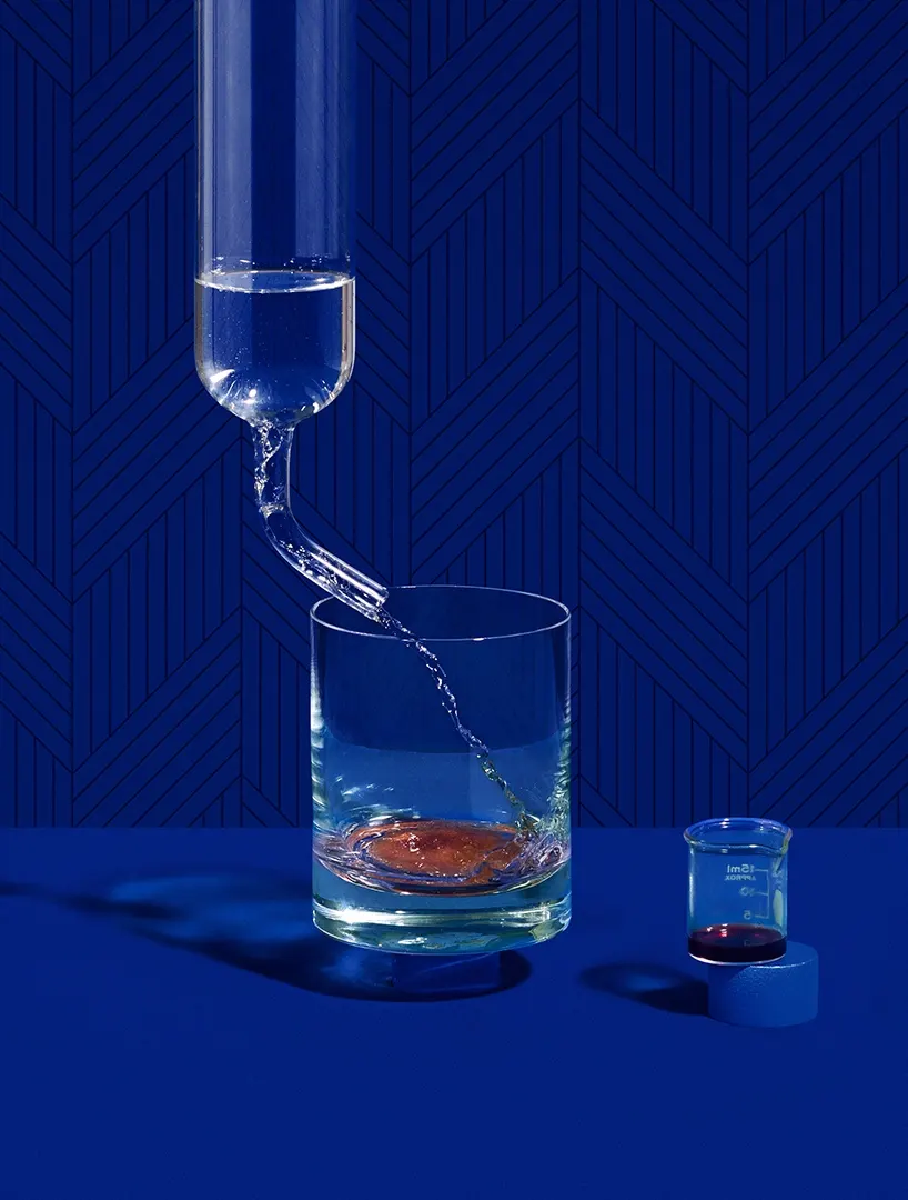 A glass is being filled with water from a glass tube on a blue surface, with a small beaker containing a dark liquid nearby. The background is a patterned blue wall.