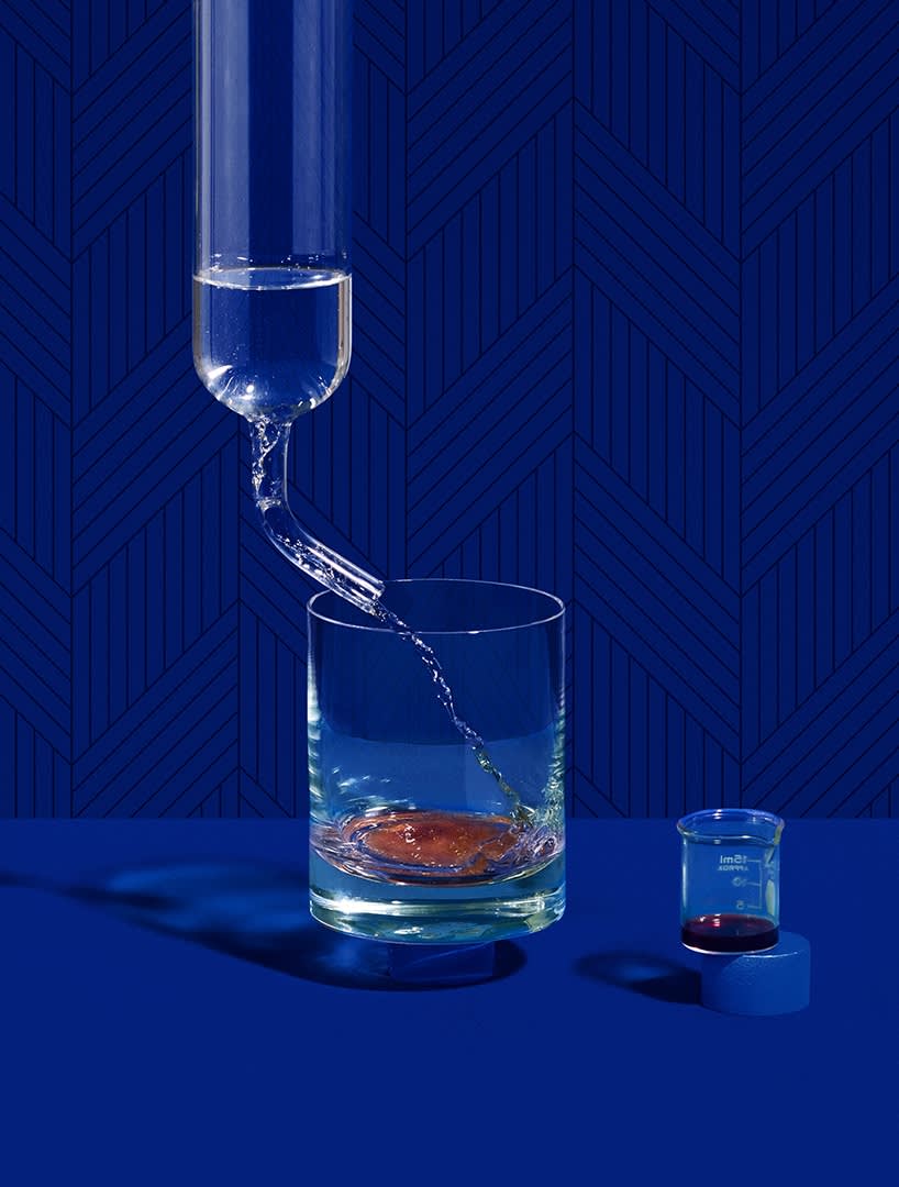 A glass is being filled with water from a glass tube on a blue surface, with a small beaker containing a dark liquid nearby. The background is a patterned blue wall.