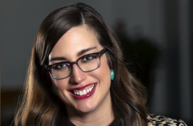 A woman with long brown hair, wearing glasses, turquoise earrings, and a printed top, smiles at the camera against a softly blurred dark background.