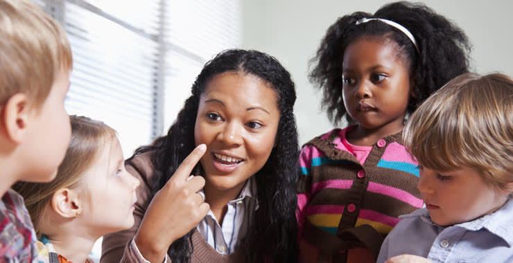 A woman is smiling and pointing to her face while talking to four young children who are attentively listening and watching her in a brightly lit room.