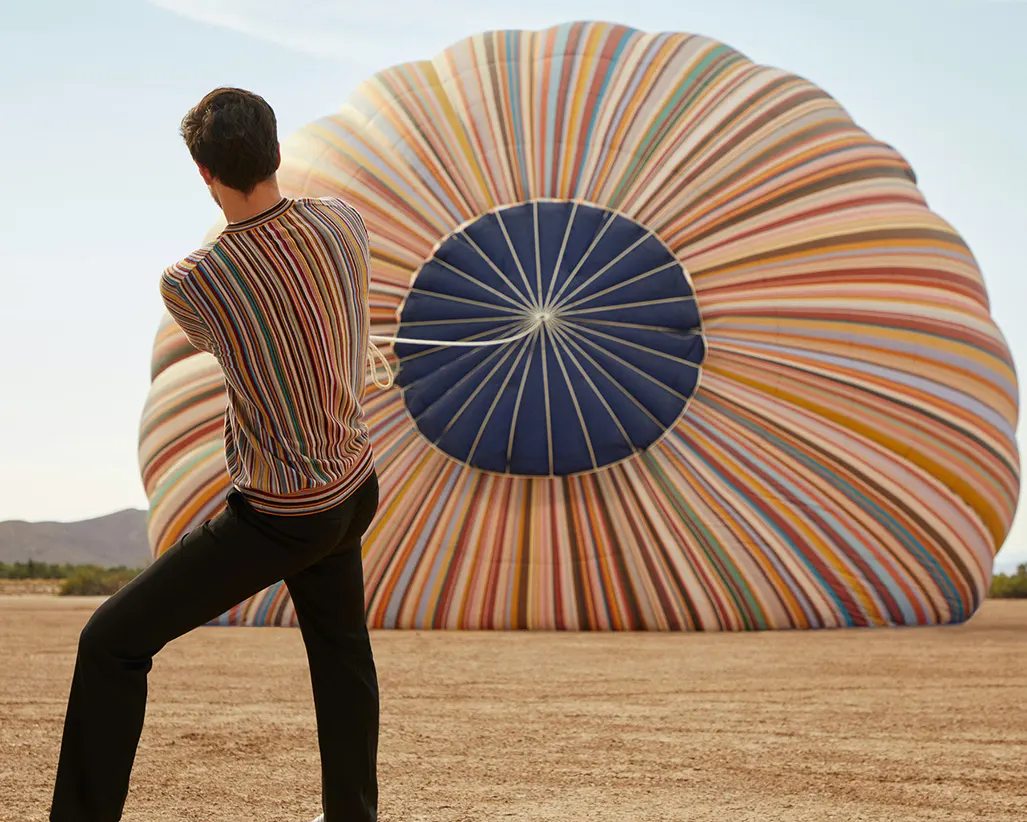 A person stands on dry ground pulling on a rope attached to a large, colorful hot air balloon lying deflated on the ground, with stripes in orange, yellow, red, blue, and white. The scene is set outdoors in daylight.