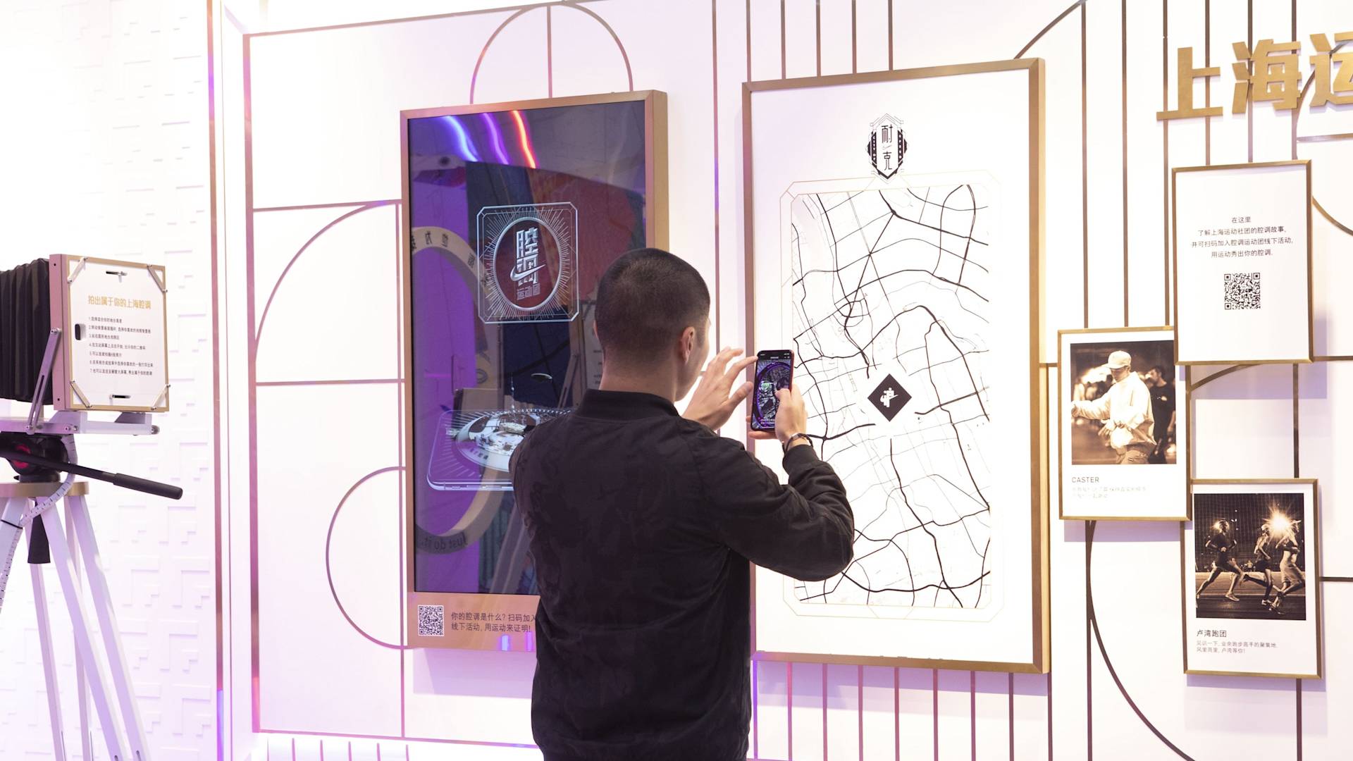 A man takes a photo of a framed map on a wall display with his phone. The wall features other framed items, a QR code, Chinese text, and a vintage camera on a tripod.