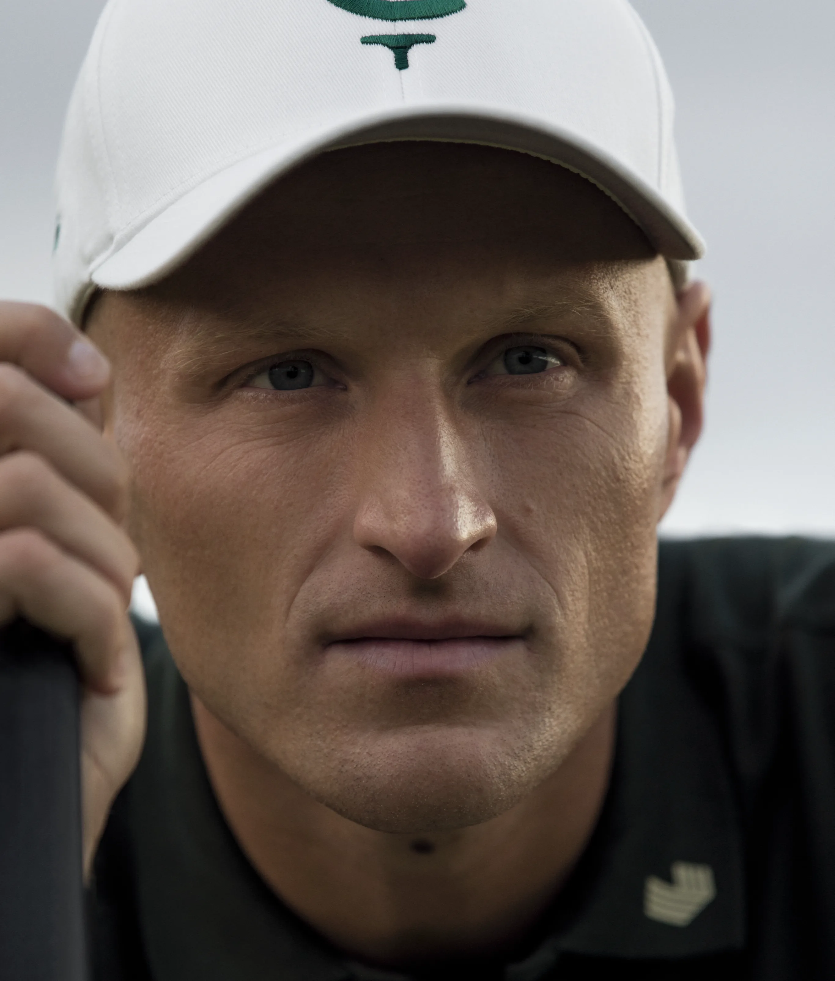A close-up of a golfer's face, wearing a white Cleeks cap and holding a golf club.