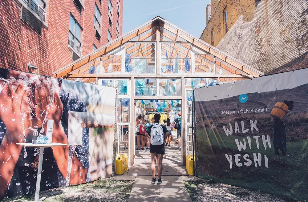 A person with a backpack enters a glass greenhouse decorated with blue and white balloons. Banners outside read “WALK WITH YESHI” and show a woman carrying a yellow water container. Several people are inside the greenhouse.