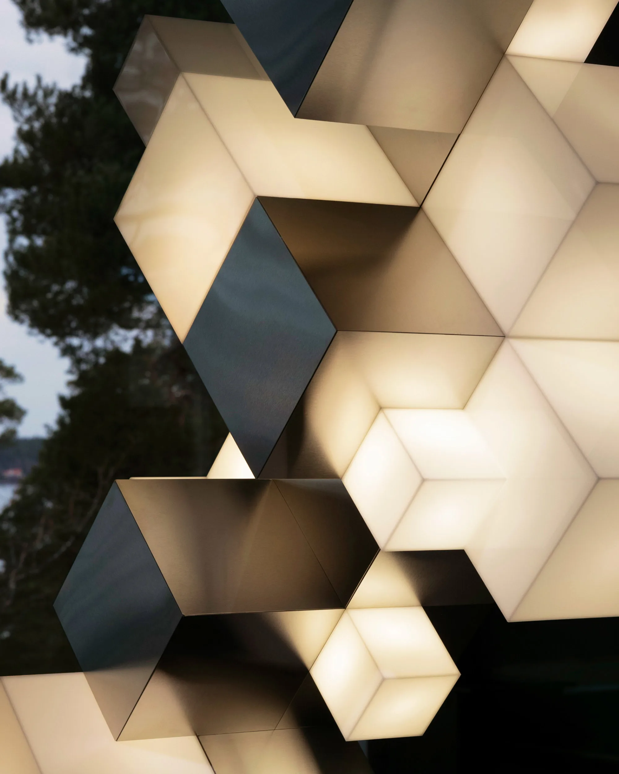 Geometric light fixtures shaped like cubes are clustered together, glowing softly with warm white light. The cubes overlap, creating a modern, abstract pattern against a background of trees.