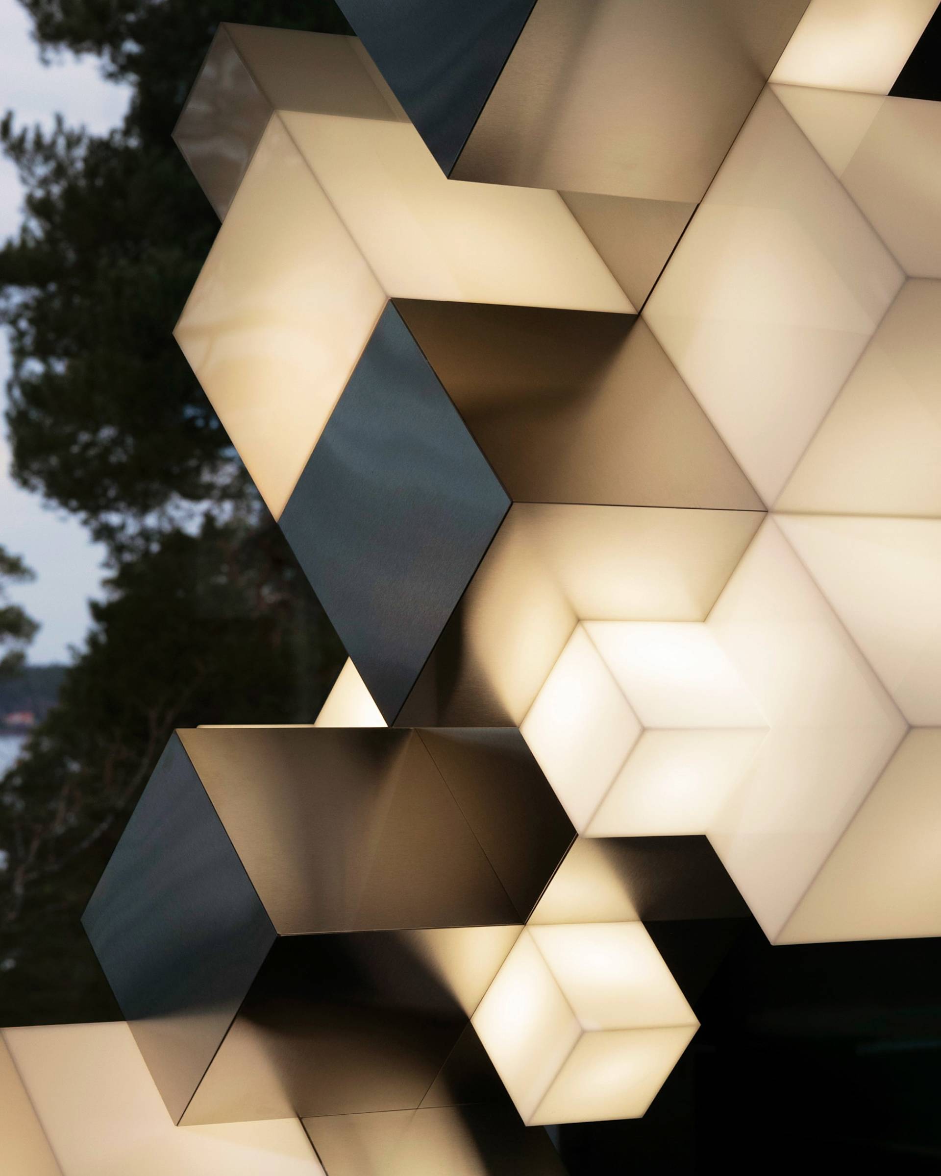 Geometric light fixtures shaped like cubes are clustered together, glowing softly with warm white light. The cubes overlap, creating a modern, abstract pattern against a background of trees.