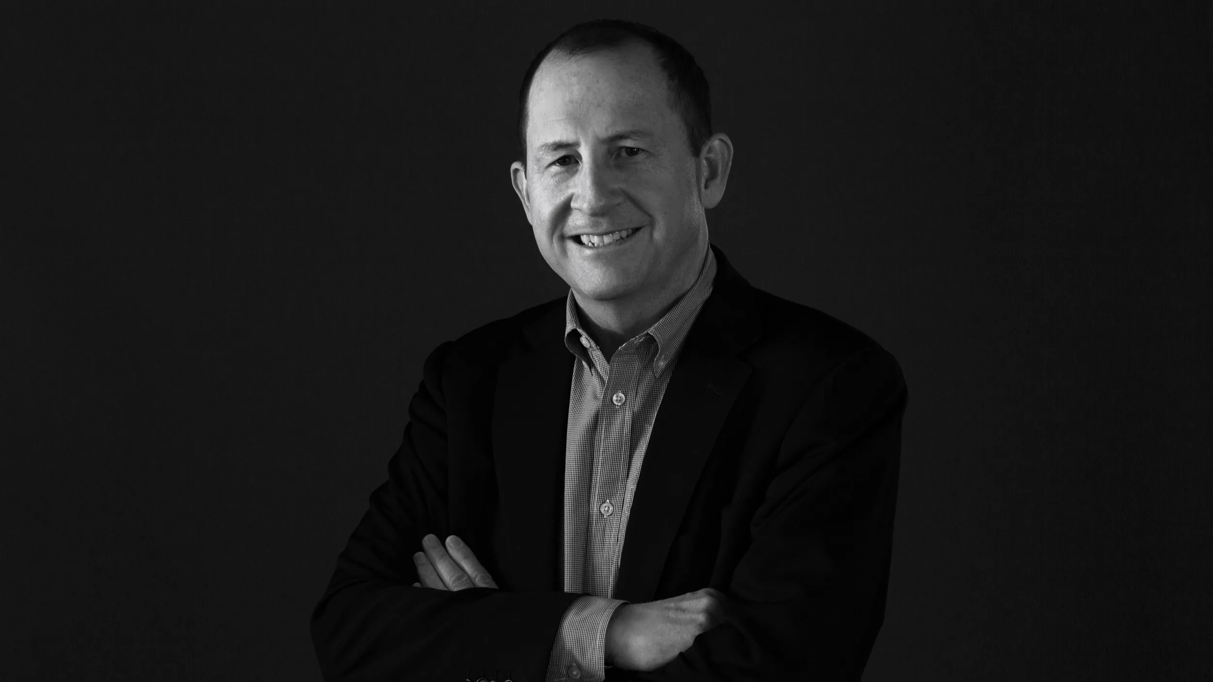 A man wearing a suit jacket and a button-down shirt stands with his arms crossed, smiling at the camera against a dark background.