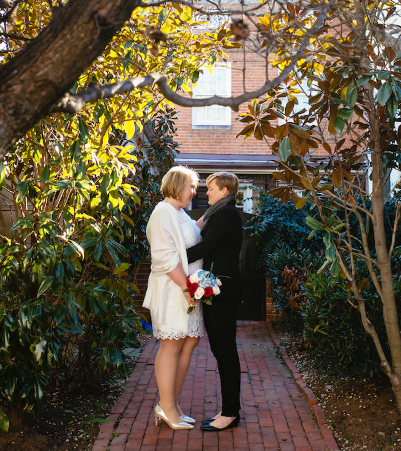 Two women stand facing each other on a brick path, surrounded by greenery and trees. One wears a white dress holding a bouquet, the other wears a black suit. They smile at each other in a sunlit garden setting.