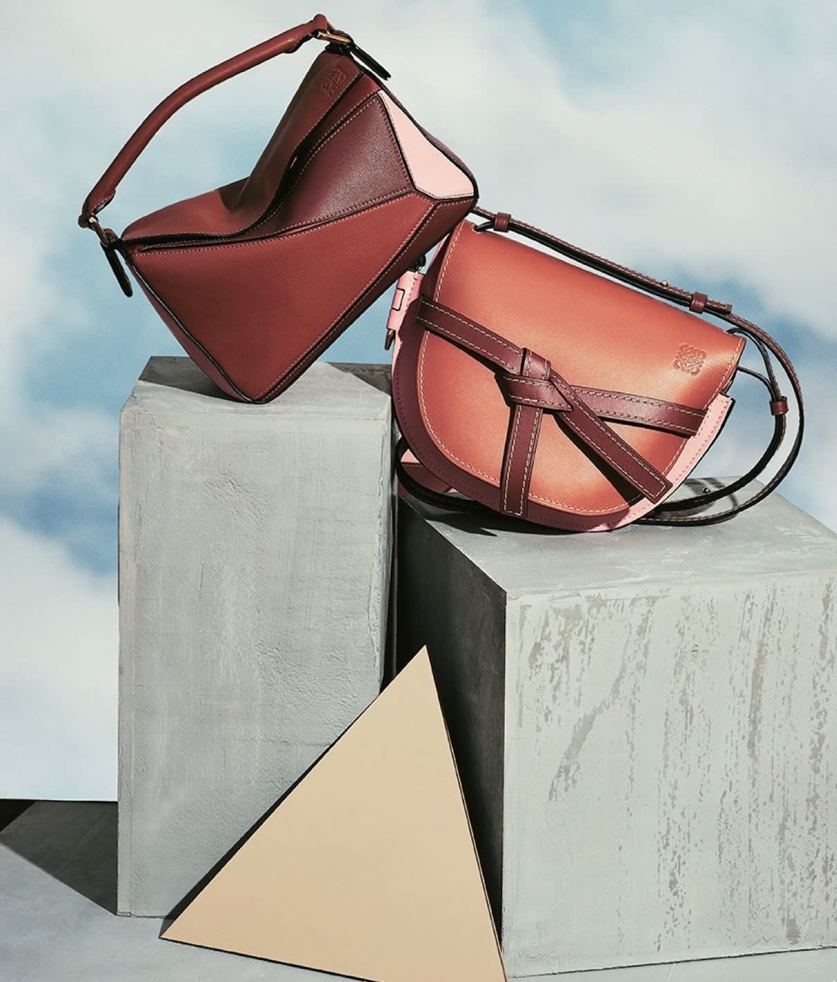Two designer handbags in shades of brown and pink are displayed on gray concrete blocks, with a beige triangular prism in front. The background is softly blurred in blue and white tones.