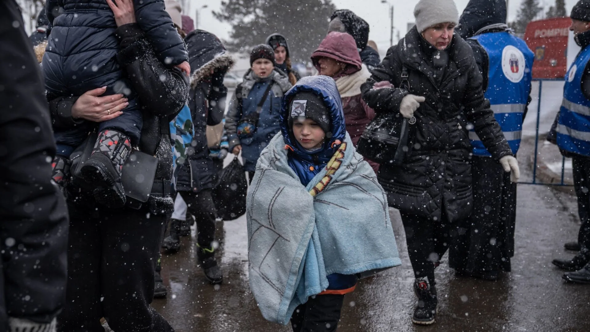 A group of people, including a child wrapped in a blanket, walk through falling snow. Most are bundled in winter clothes, and some adults wear blue vests. The scene appears cold and somber.