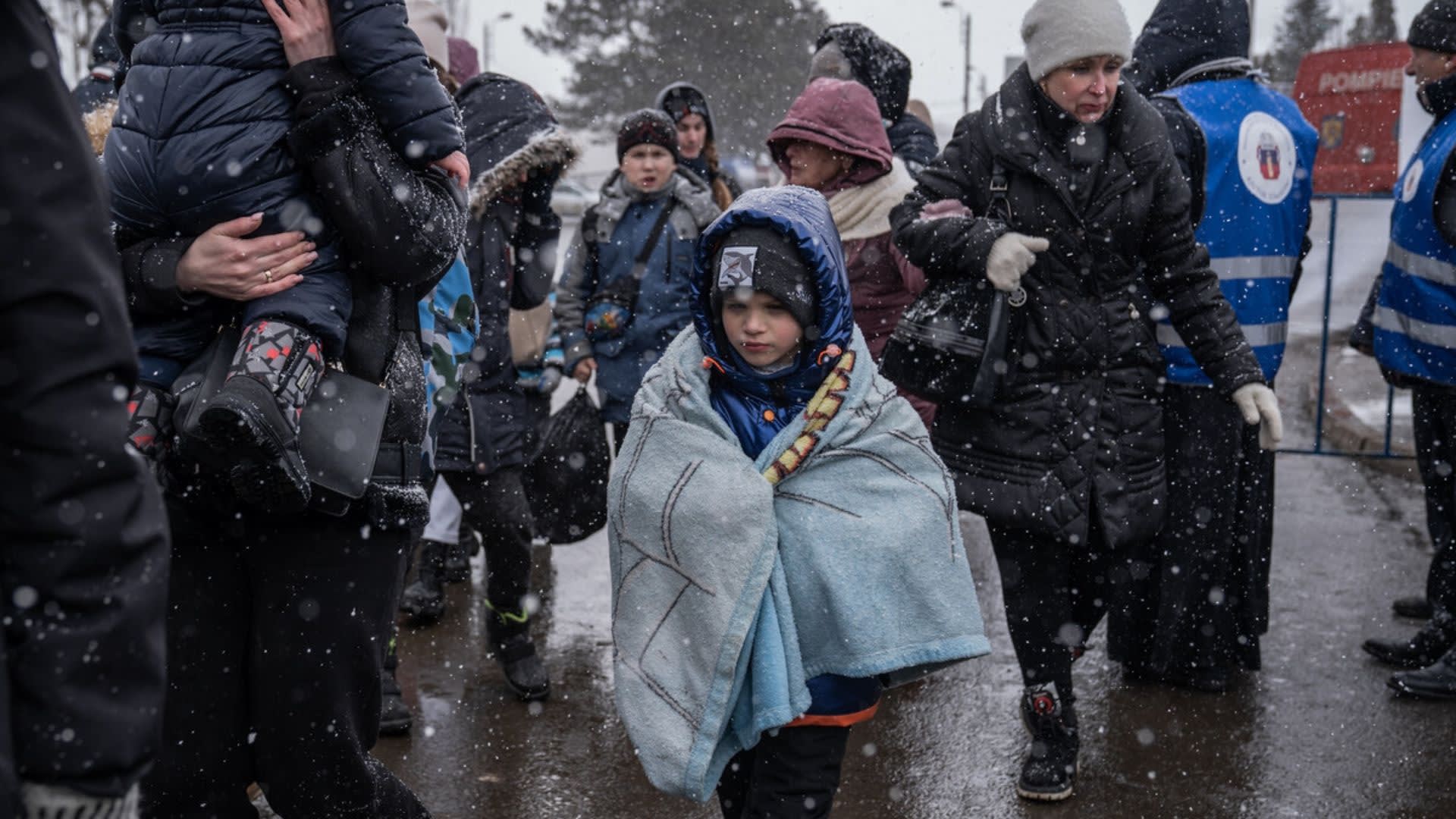 A group of people, including a child wrapped in a blanket, walk through falling snow. Most are bundled in winter clothes, and some adults wear blue vests. The scene appears cold and somber.
