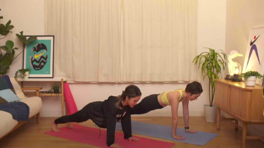 Two women practice yoga indoors on mats, both holding a plank pose. The room is cozy, with plants, a sofa, framed art, and soft lighting. One woman wears black, the other wears a yellow sports bra and gray leggings.