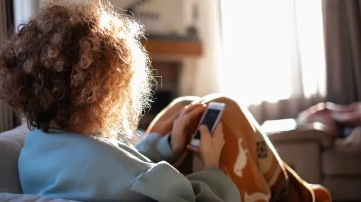 A person with curly hair sits on a couch in a sunlit room, holding a smartphone and wearing a cozy blanket with an animal pattern. The scene appears relaxed and peaceful.