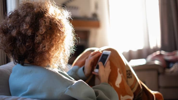 A person with curly hair sits on a couch in a sunlit room, holding a smartphone and wearing a cozy blanket with an animal pattern. The scene appears relaxed and peaceful.