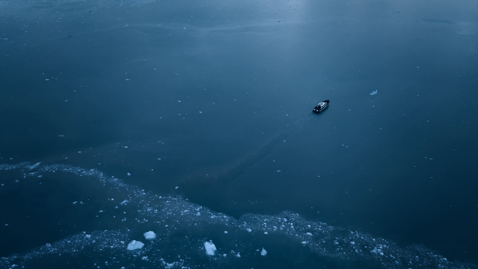 A small boat floats alone on a vast, icy blue body of water, surrounded by scattered ice chunks and leaving a faint trail behind.