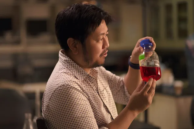 A man with dark hair and a patterned shirt holds up and examines a laboratory flask filled with red liquid in a lab setting.