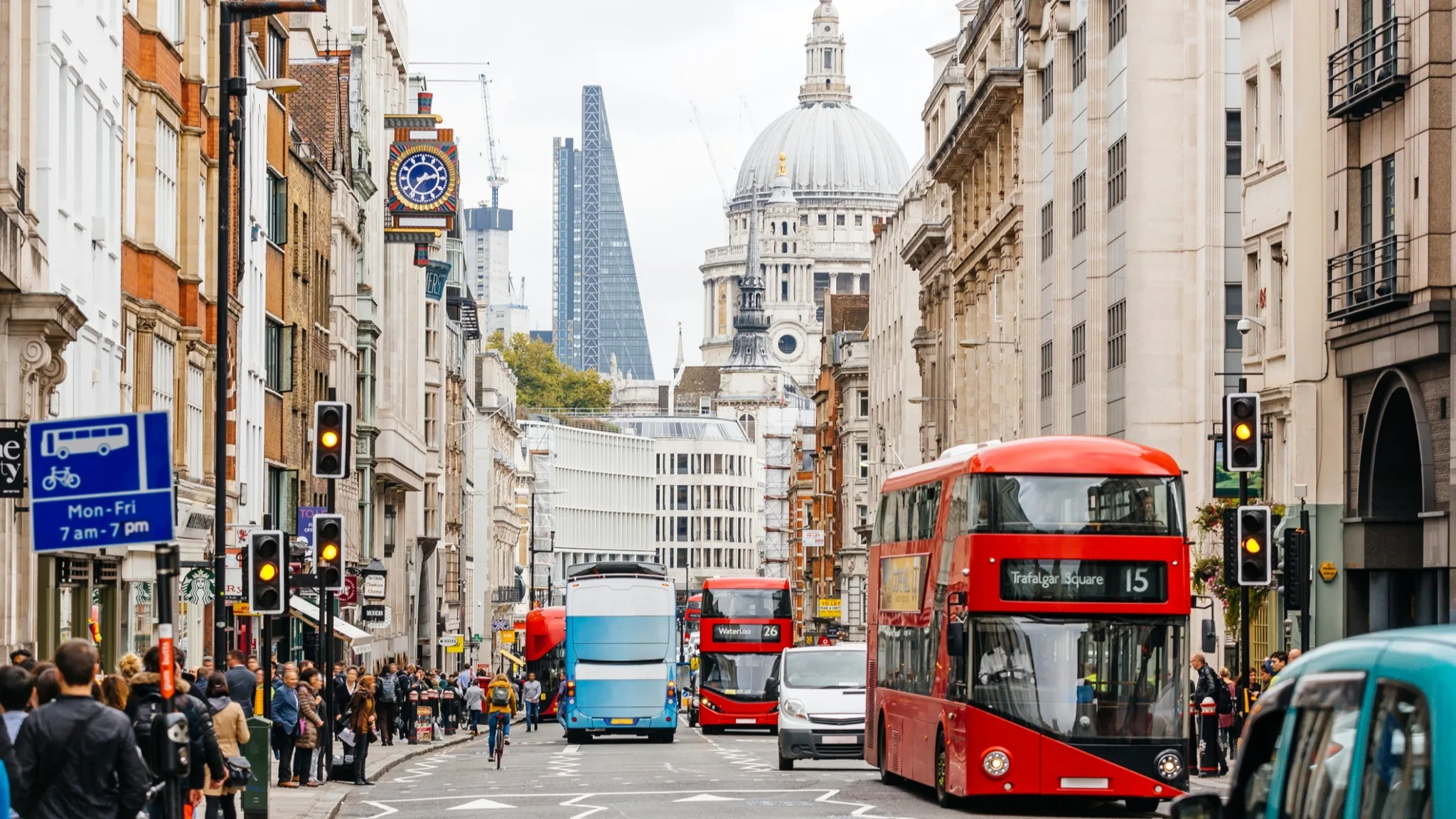 Busy London street with red double-decker buses, pedestrians on sidewalks, and traffic lights. St. Paul’s Cathedral and modern skyscrapers are visible in the background under a cloudy sky.