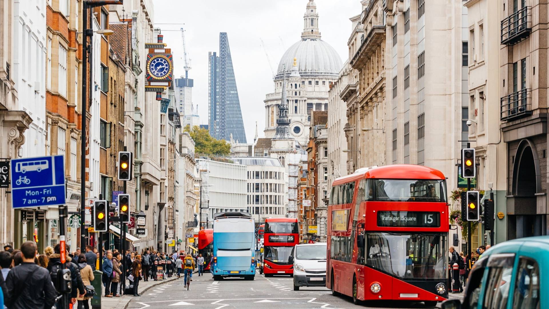 Busy London street with red double-decker buses, pedestrians on sidewalks, and traffic lights. St. Paul’s Cathedral and modern skyscrapers are visible in the background under a cloudy sky.