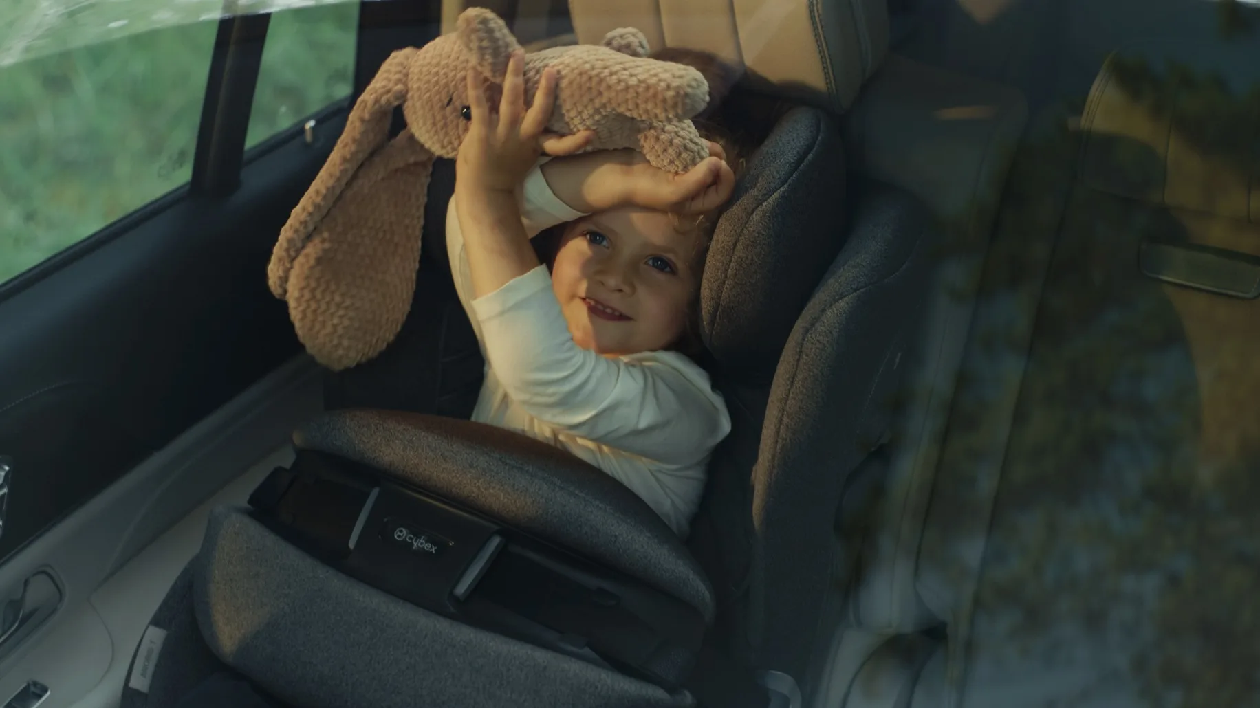 A young child sits in a car seat, smiling and holding a plush bunny toy above their head, viewed through a car window.