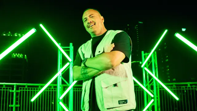A man wearing a light vest and dark shirt stands with arms crossed, smiling, in front of green neon lights and outdoor railings at night, with city buildings in the background.