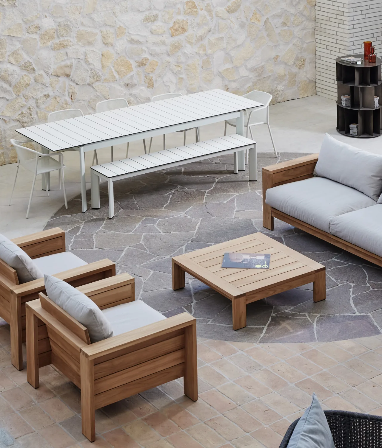 Modern outdoor patio with a wooden lounge set, gray cushions, a square coffee table with magazines, and a long white dining table with benches and chairs, set on a stone floor against a light stone wall.