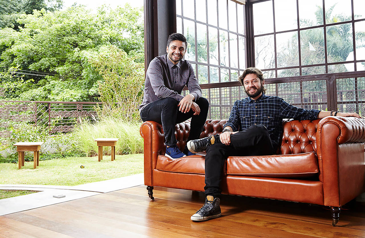 Two men indoors by large windows, one sitting on a brown leather sofa and the other perched on its armrest, smiling. Lush greenery and wooden benches are visible outside through the windows.