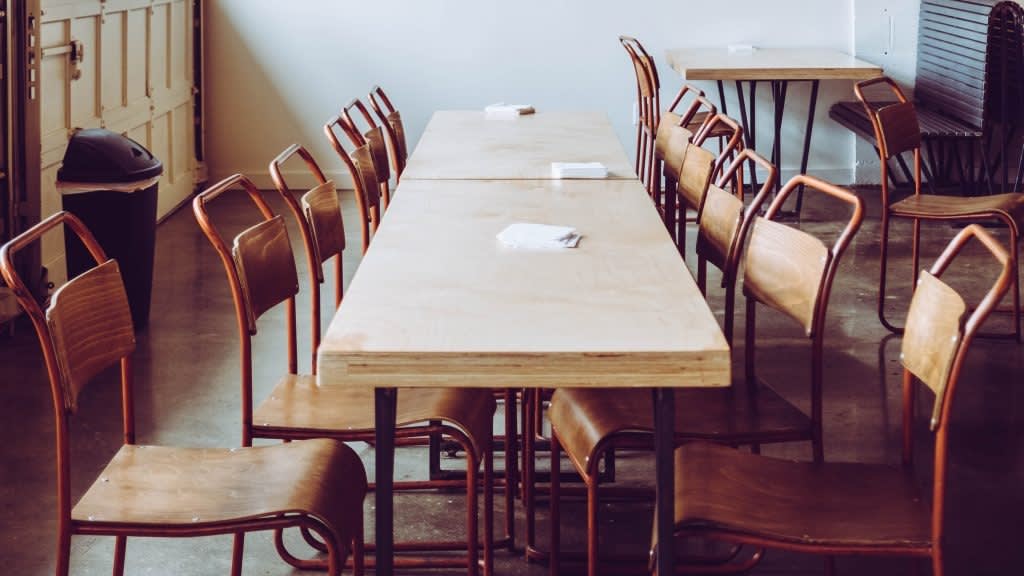 Two wooden tables pushed together with several wooden chairs around them in an empty room. Each table has small stacks of napkins placed on top. The setting appears quiet and unoccupied.