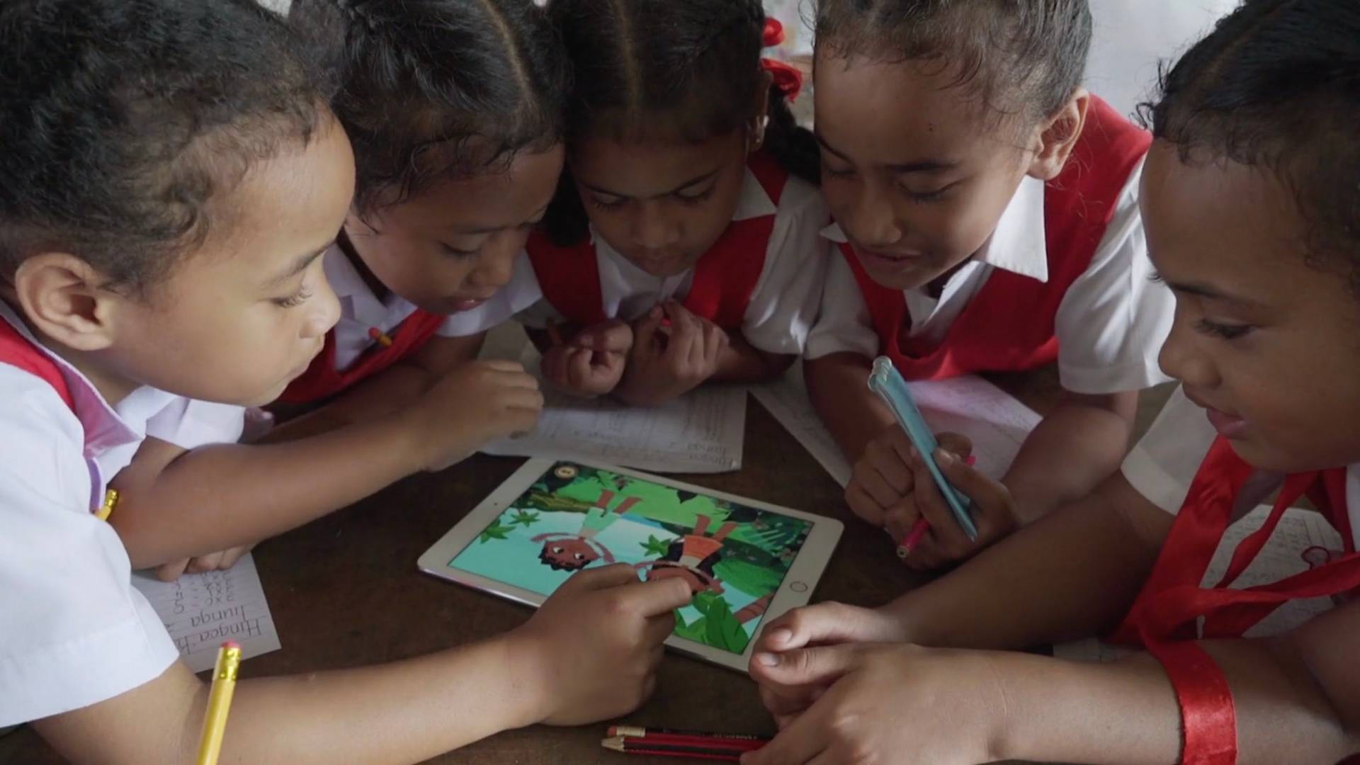 Five young children in red and white school uniforms are gathered around a tablet, interacting with an educational game, while holding pens and looking focused and engaged.