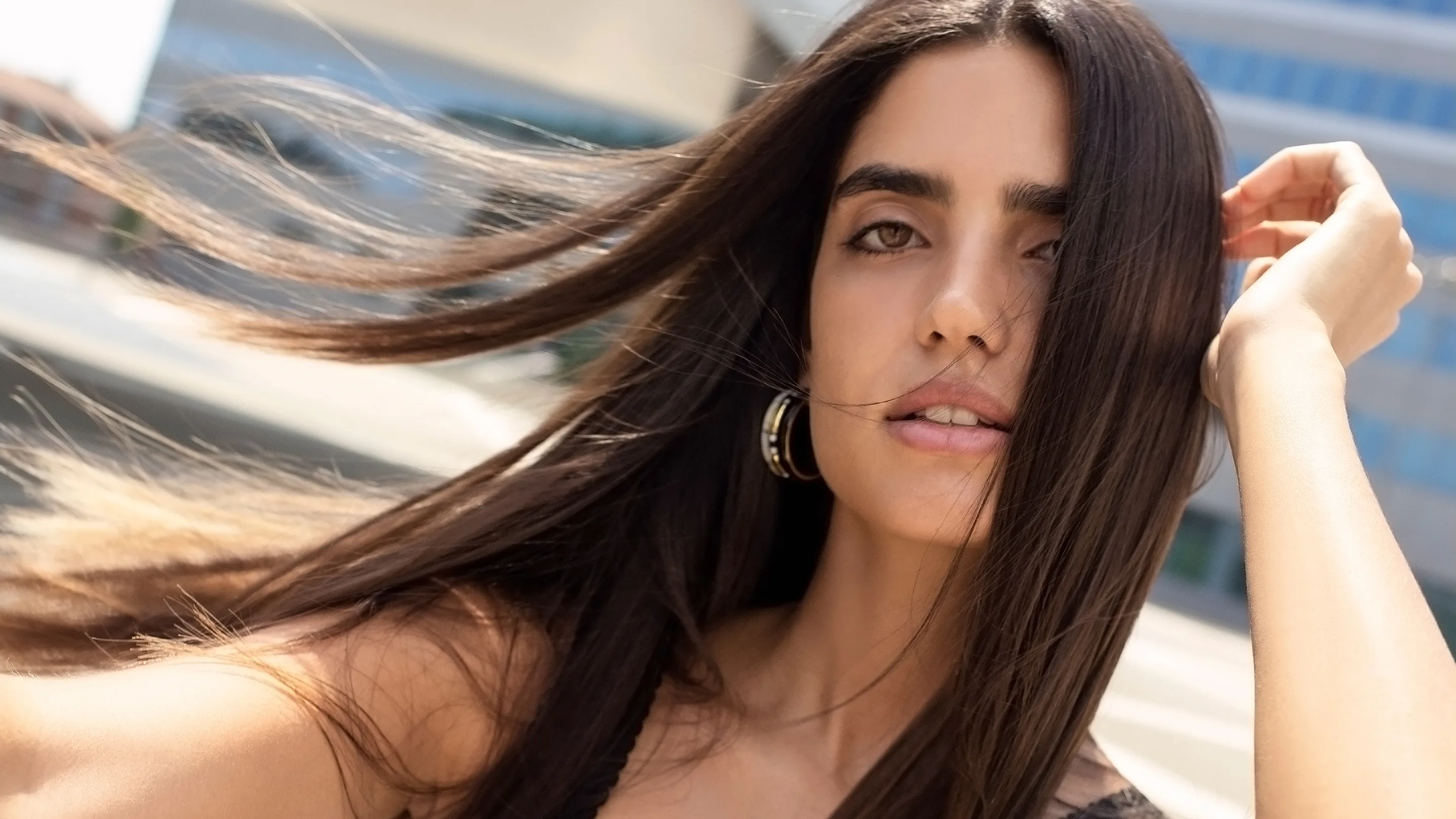 A young woman with long, dark hair poses outdoors, her hair blowing in the wind. She looks directly at the camera with a soft expression, wearing hoop earrings and a sleeveless top, with blurred buildings in the background.